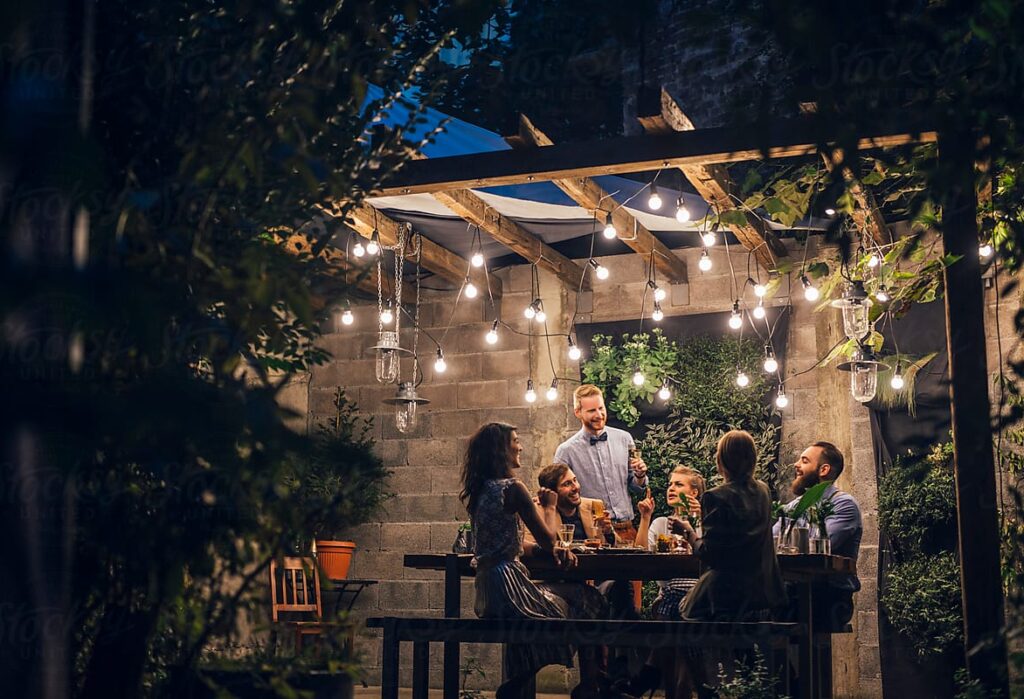 “Group of adults enjoying a cozy evening in a beautifully landscaped backyard with warm string lights.”